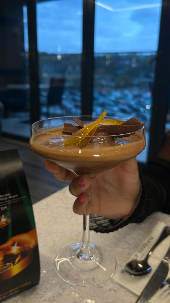 Hand holds a stemmed coupe of chocolate mousse with candied orange above a lace tablecloth and evening city lights
